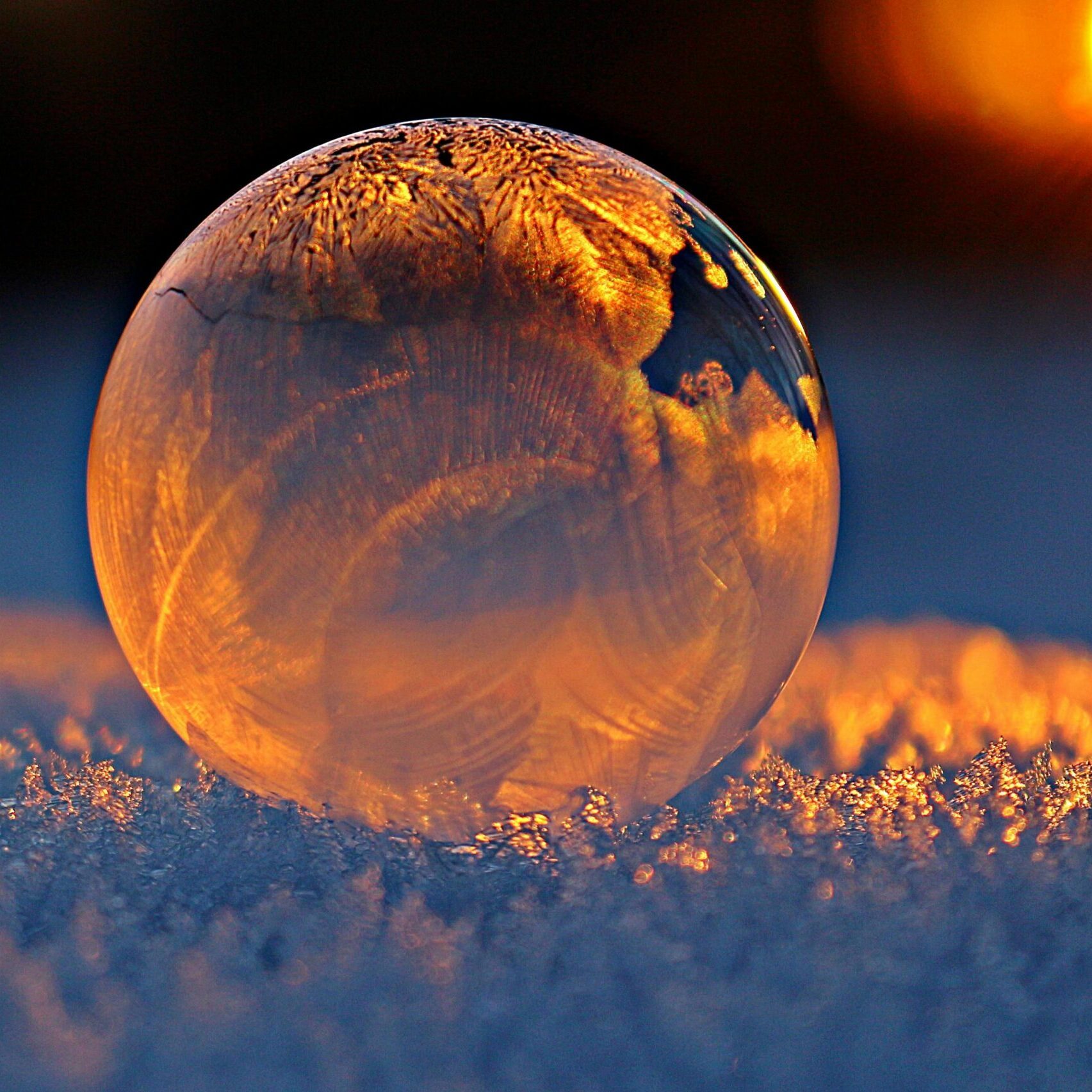 Close-up shot of a frozen bubble with warm reflections resting on a snowy surface at twilight.